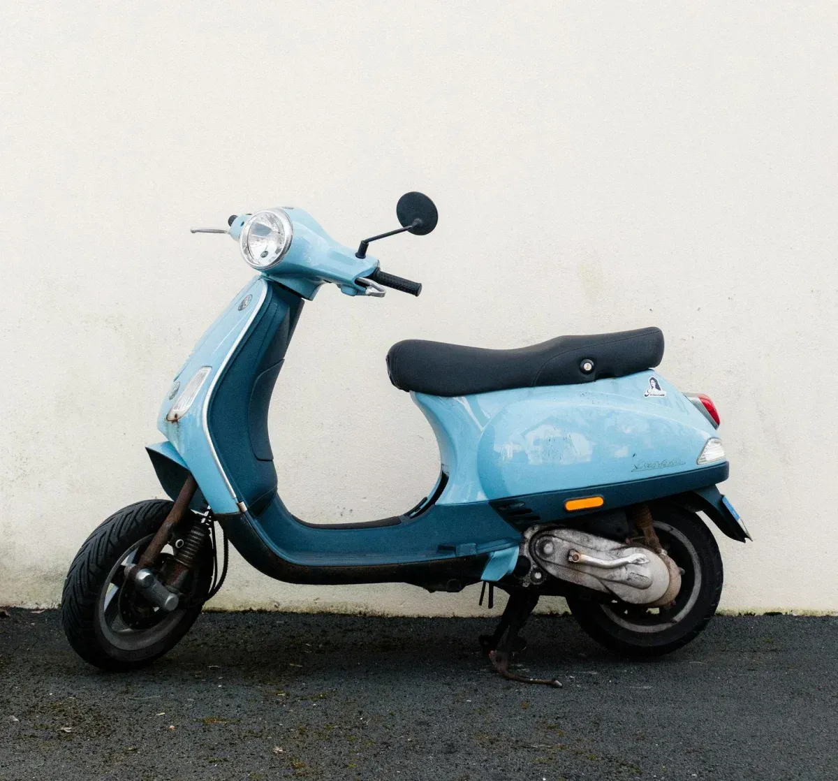 Scooter azul claro estacionado contra una pared blanca.