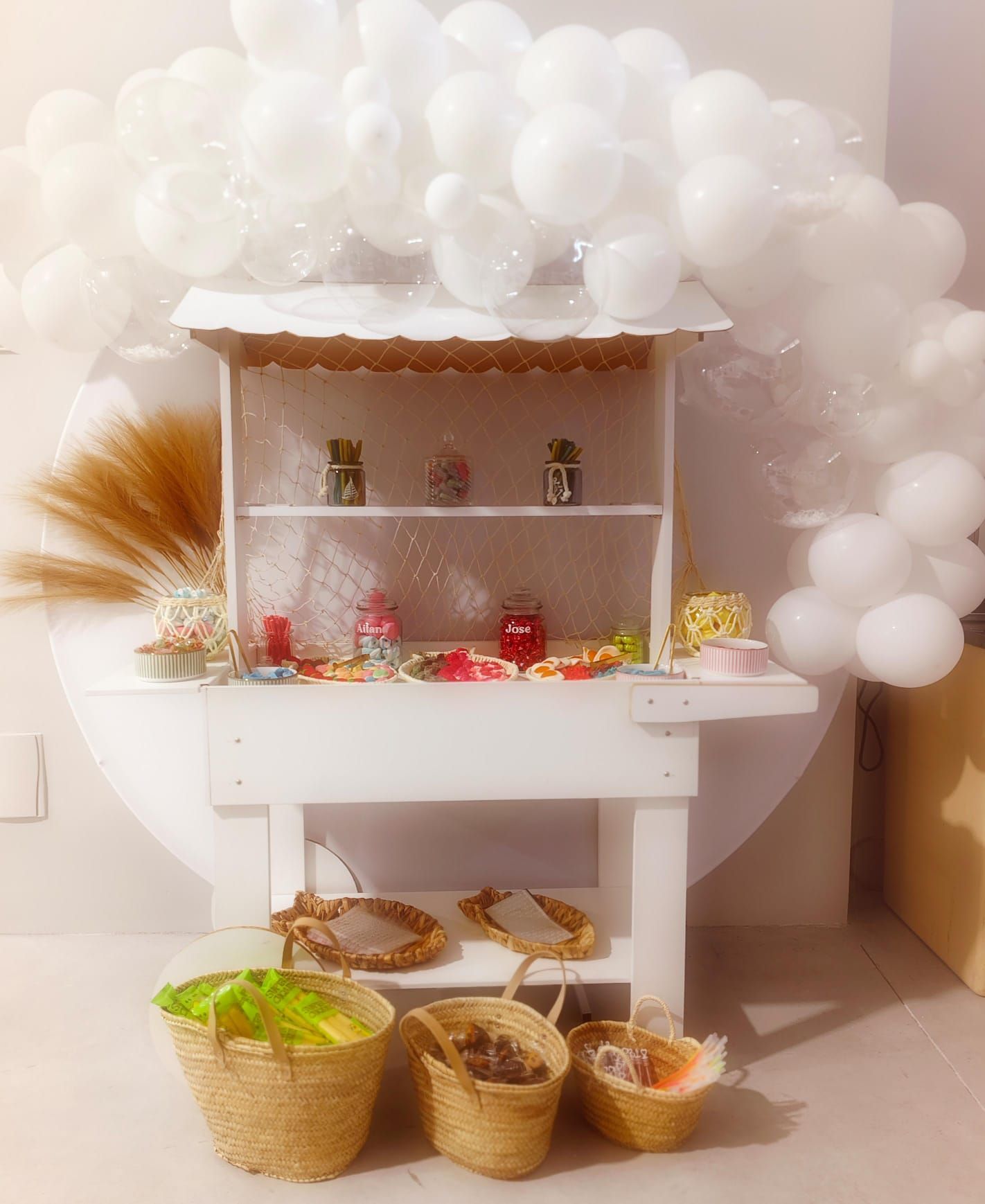 Un carrito blanco lleno de dulces y cestas está rodeado de globos blancos.