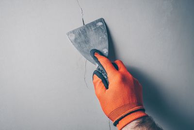 Eine behandschuhte Hand kratzt mit einem Spachtel Farbe von einer grauen Wand.