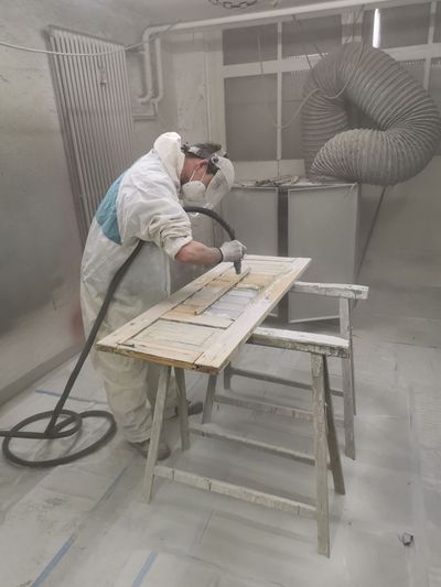 Eine Person in einem weissen Overall besprüht in einer staubigen Werkstatt ein Holzbrett mit Farbe.