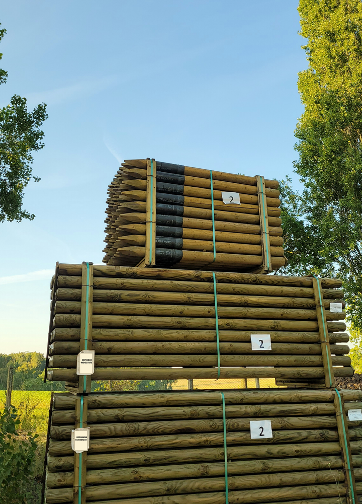 Des piles de poteaux de clôture en bois traité, empilées à l'extérieur et liées par une corde verte, se détachent sur un ciel bleu.