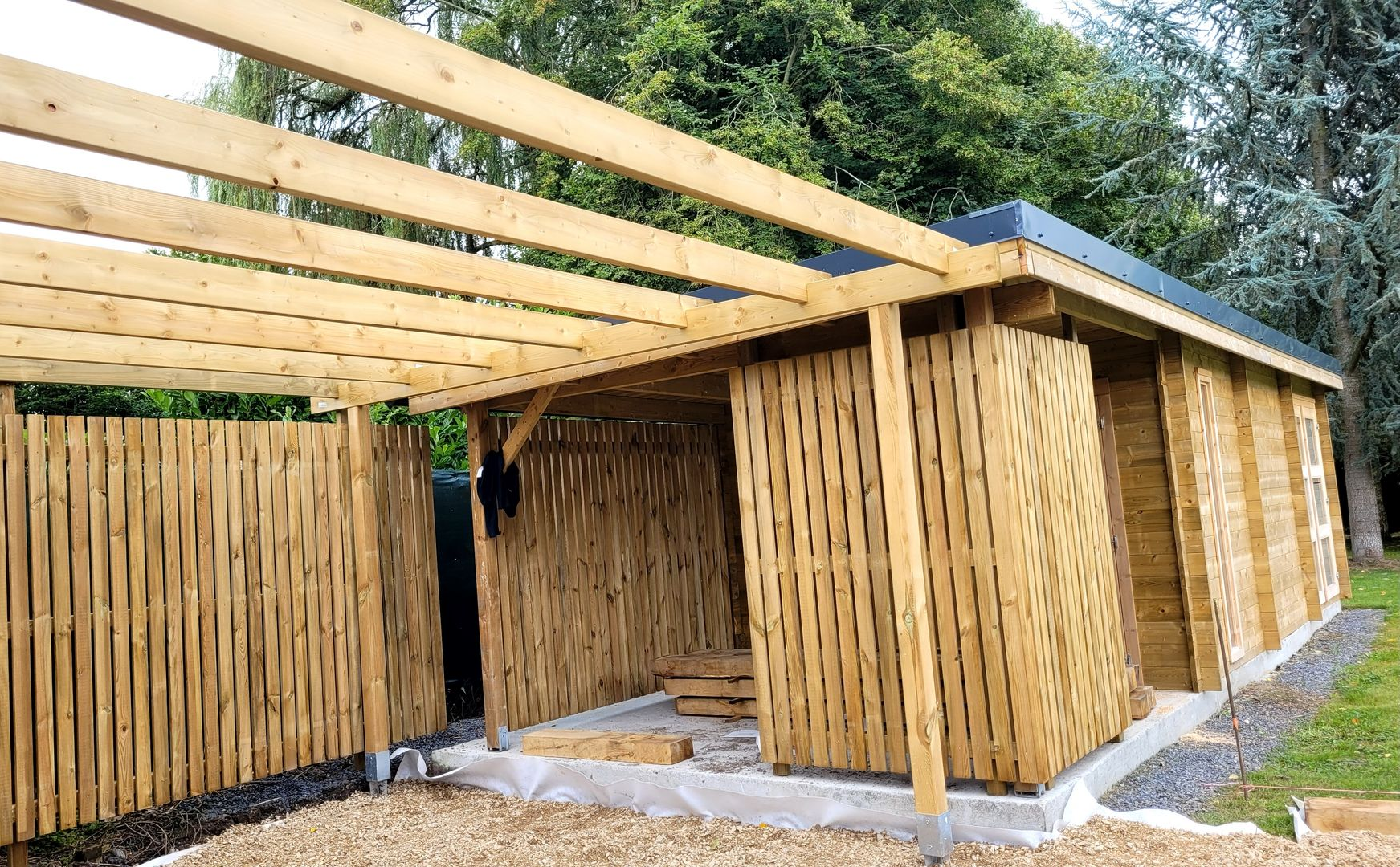 Structure en bois aux murs et au toit à claire-voie, à ciel ouvert, au bord d'un chemin de gravier.