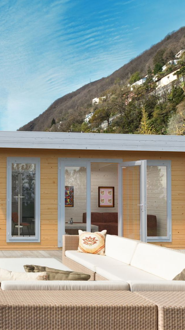 Cabane en bois sur une terrasse surplombant un lac et une montagne, avec un canapé et des fenêtres.