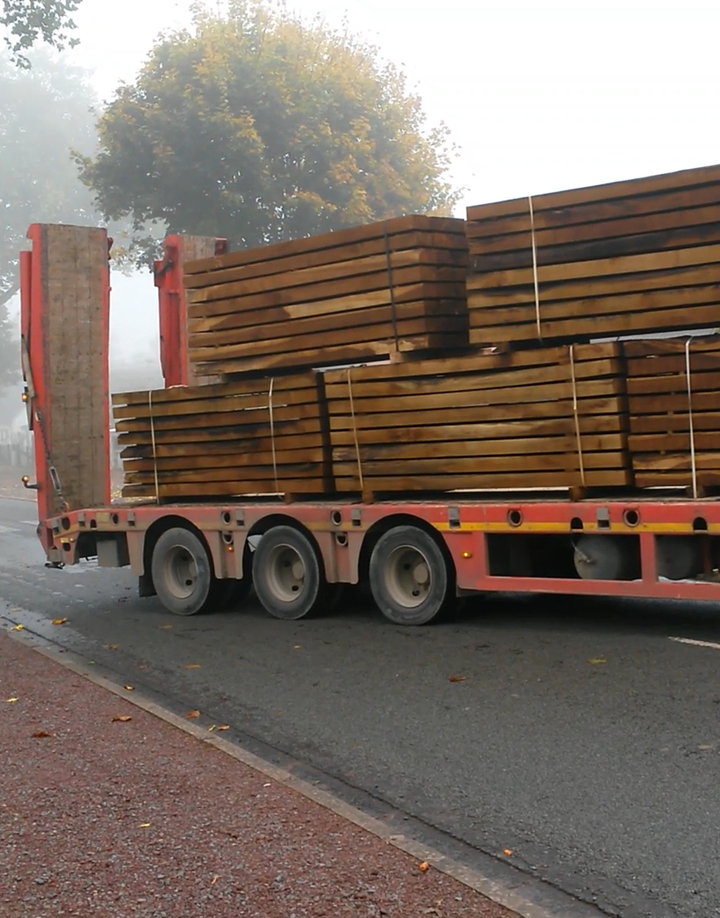 Un camion plateau rouge transporte des piles de poutres en bois sur une route brumeuse.