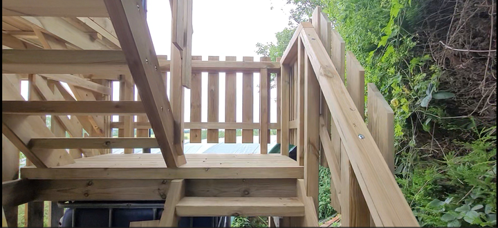 Un escalier en bois mène à un portail et à une zone verdoyante envahie par la végétation.