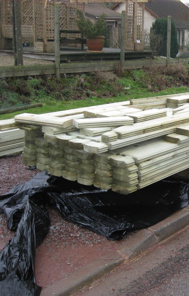 Un tas de panneaux de clôture en bois sur une bâche noire, près d'un trottoir, dans un quartier résidentiel.