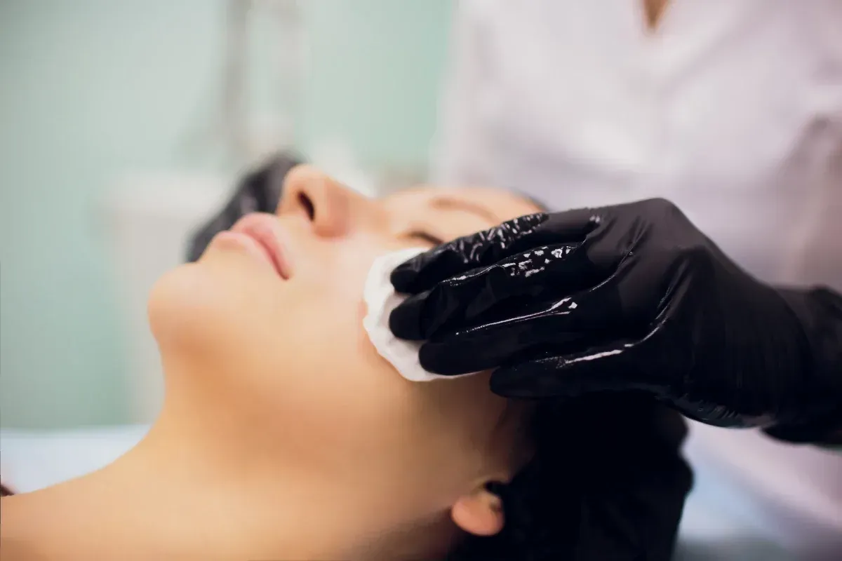 Persona recibiendo tratamiento facial; mano enguantada sosteniendo un disco de algodón contra la mejilla.