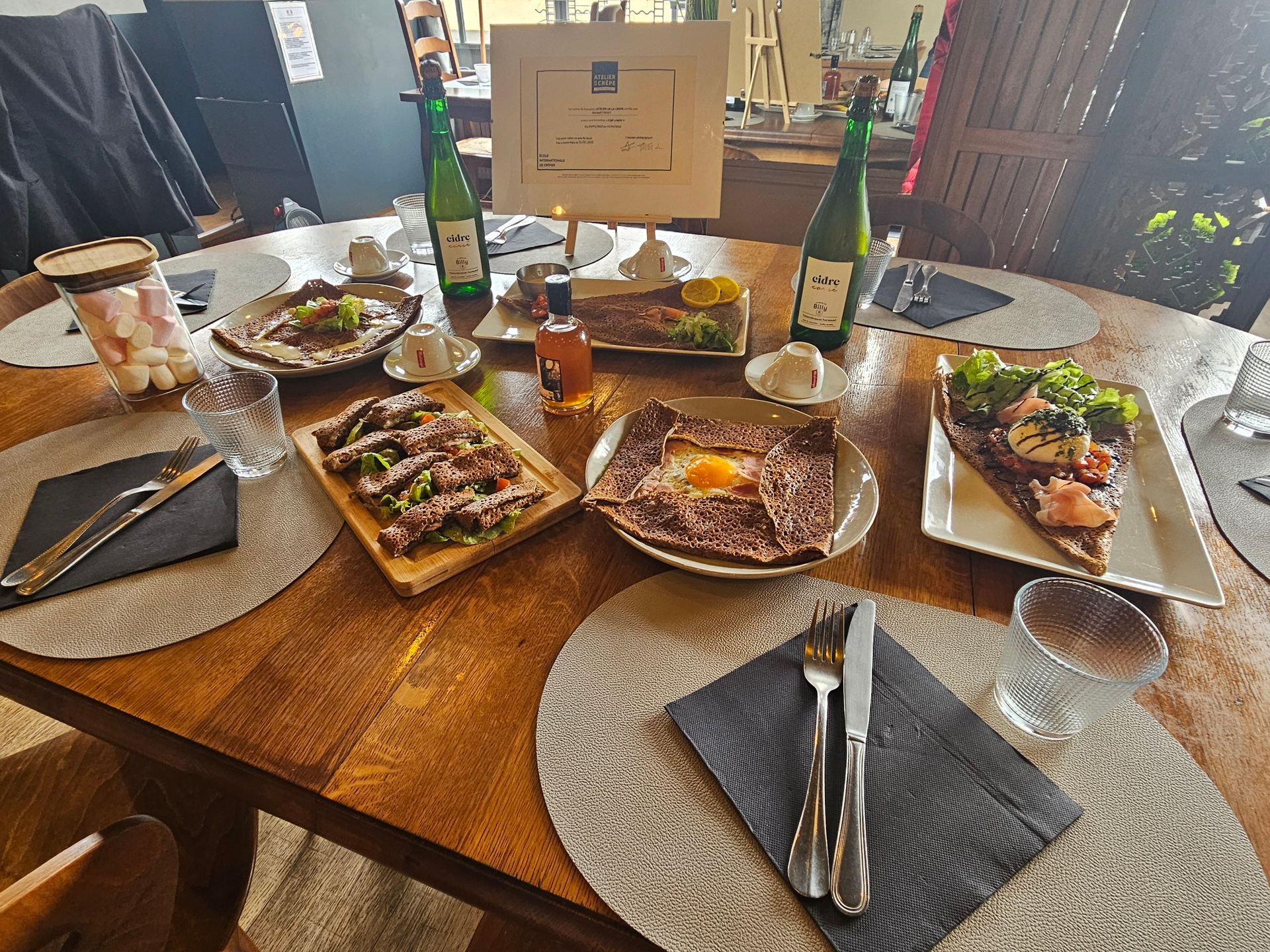 Table de restaurant dressée avec plats de galettes bretonnes salées et bouteille de cidre