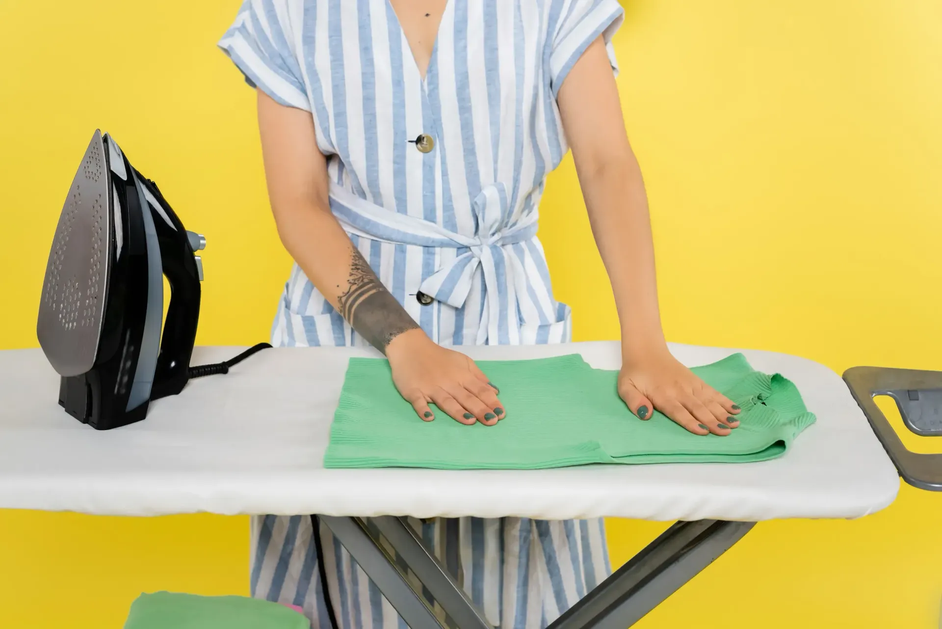 Mujer planchando una camisa verde en una tabla de planchar con una plancha gris; fondo amarillo.