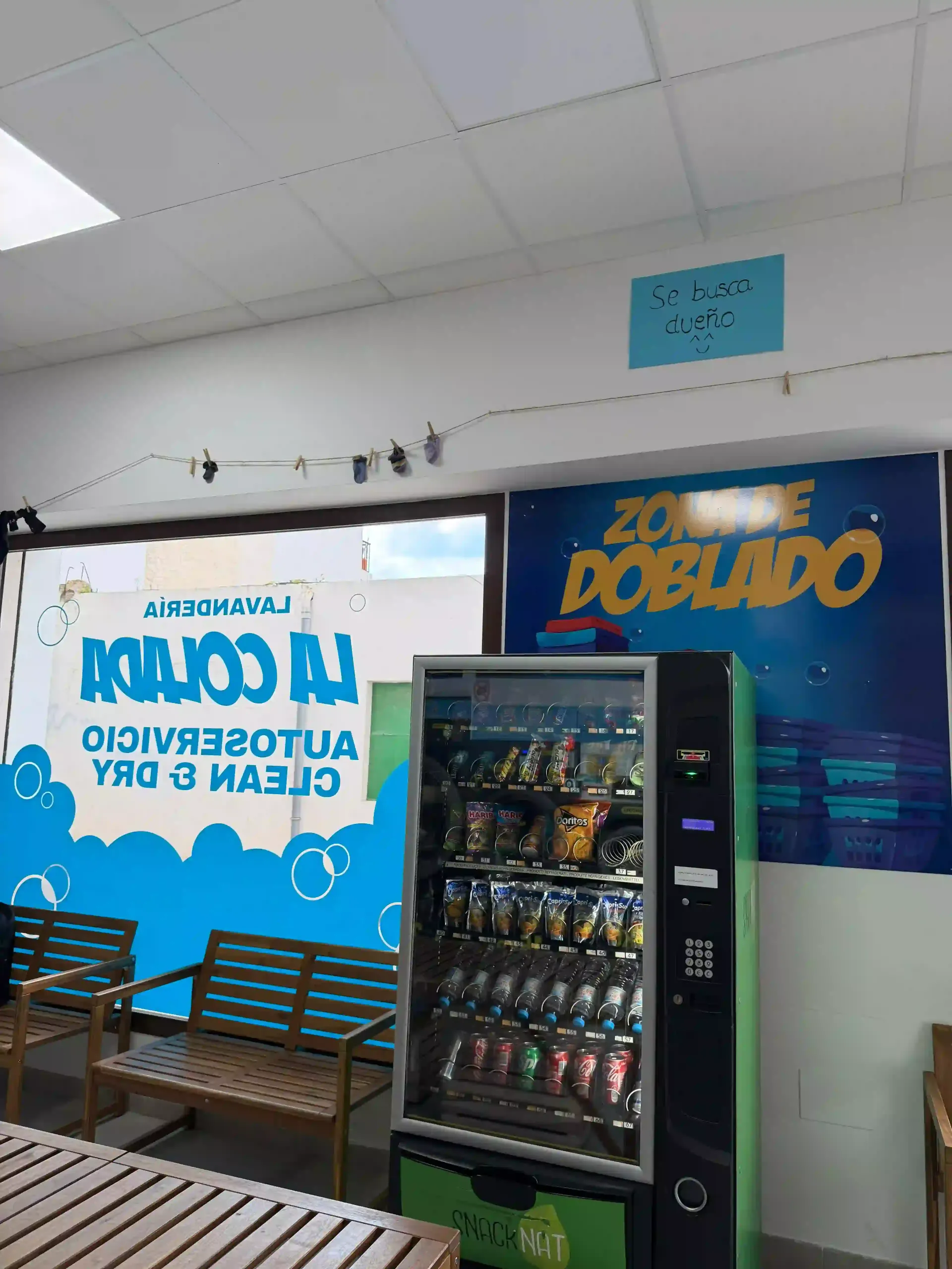Máquina expendedora en una lavandería, junto a una ventana y un banco. Letrero de 