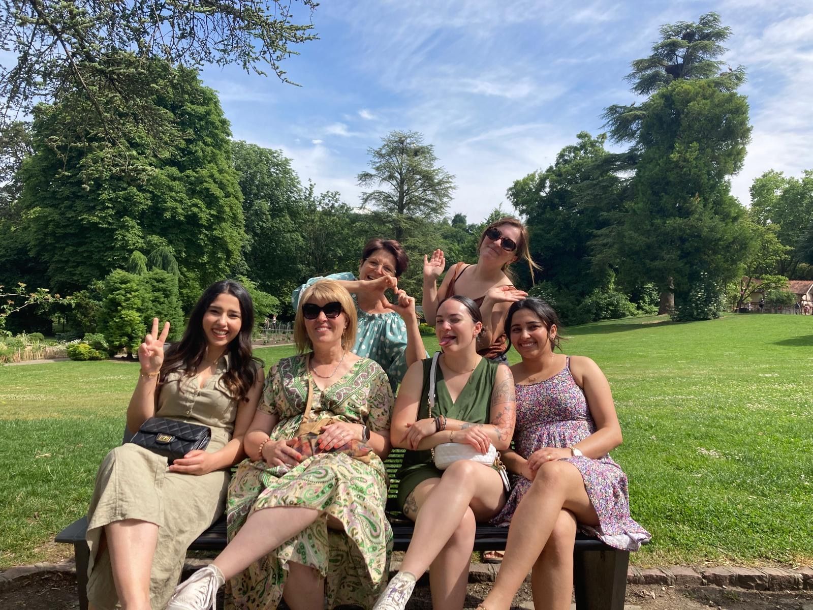 L'équipe de Coiffurama assise sur un banc dans un parc, sous un ciel bleu, souriant et faisant des signes de la main. 