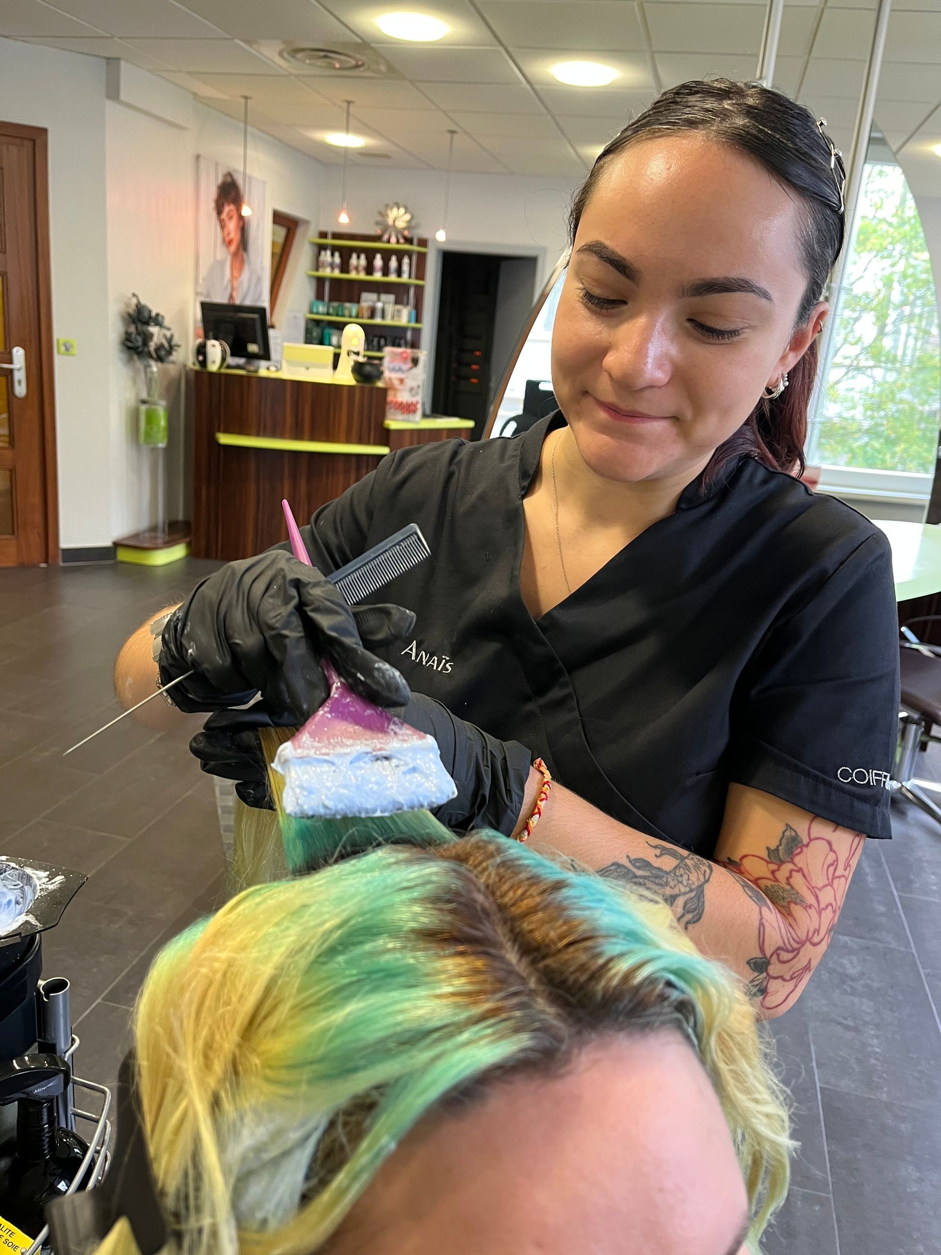 Une coiffeuse applique une teinture sur des cheveux dans un salon. Elle porte des gants, tient un pinceau, et les cheveux présentent des reflets verts et jaunes.