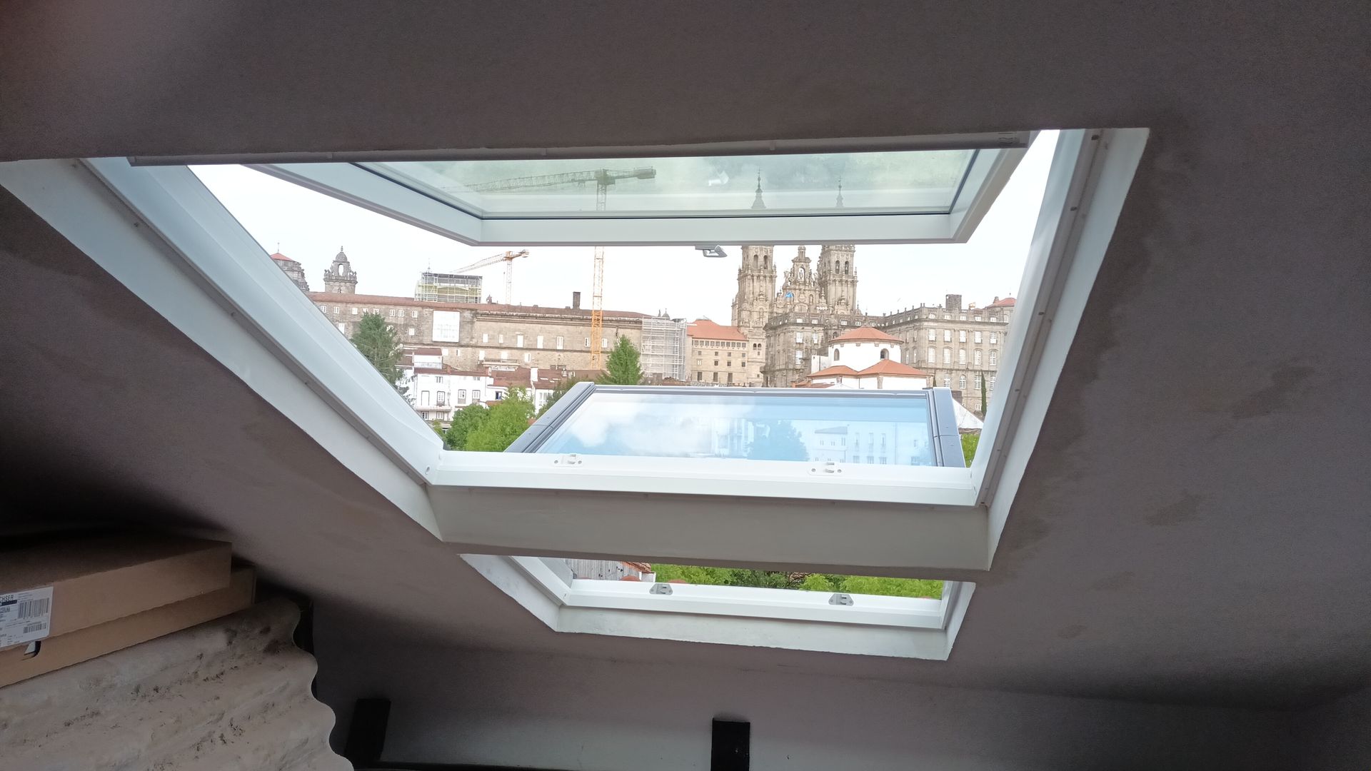 Tragaluz abierto en el ático, que muestra una vista del paisaje urbano con edificios beige y cielo despejado.