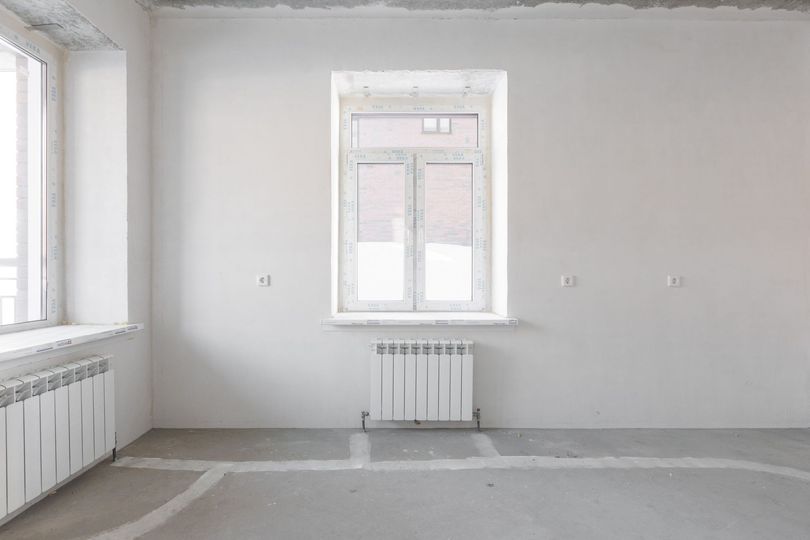 Habitación vacía, de paredes blancas, con dos ventanas, un radiador y suelo de cemento.