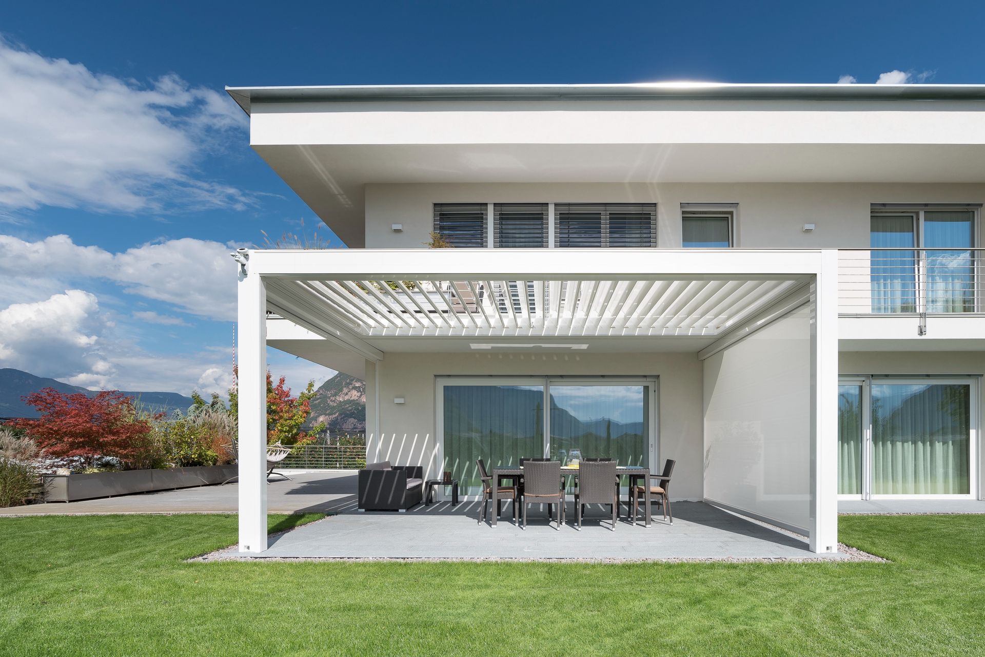 Una casa blanca con una pérgola sobre un patio con mesa y sillas.