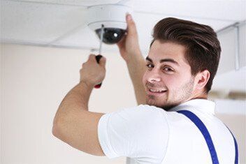 Un homme installe une caméra de sécurité au plafond et lui sourit. Il porte une chemise blanche et des bretelles bleues.