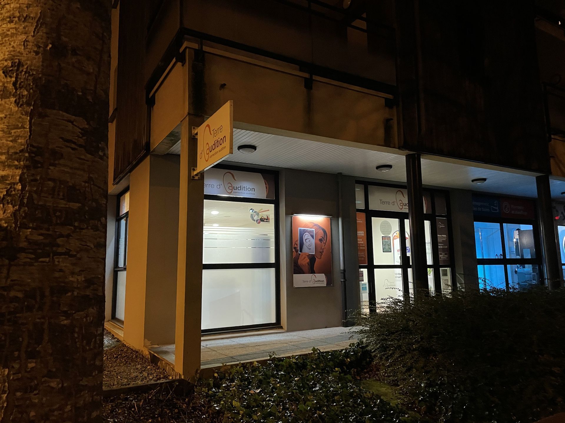 Façade de magasin nocturne avec entrée éclairée, auvent et fenêtres éclairées en bleu, donnant sur une rue étroite.