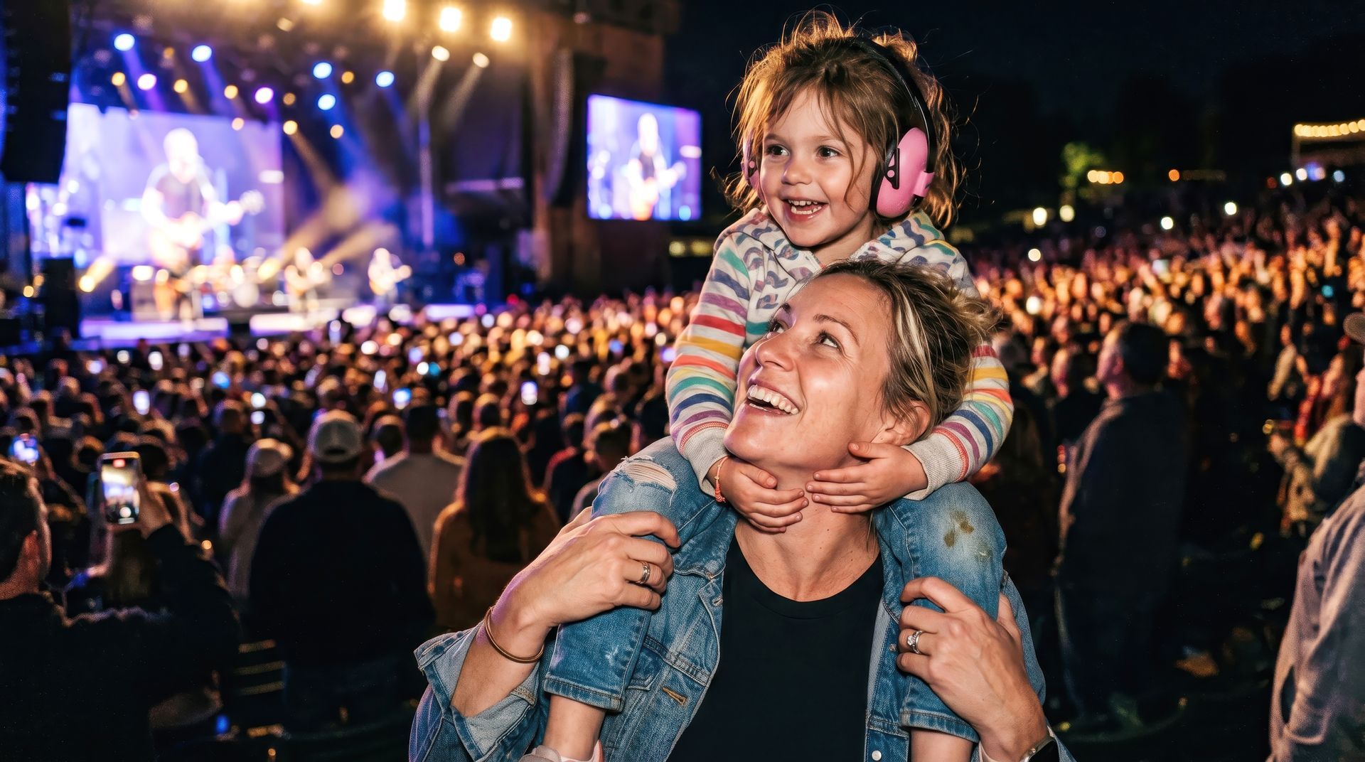 Un enfant souriant sur les épaules d'un homme lors d'un concert nocturne bondé, sous les projecteurs.