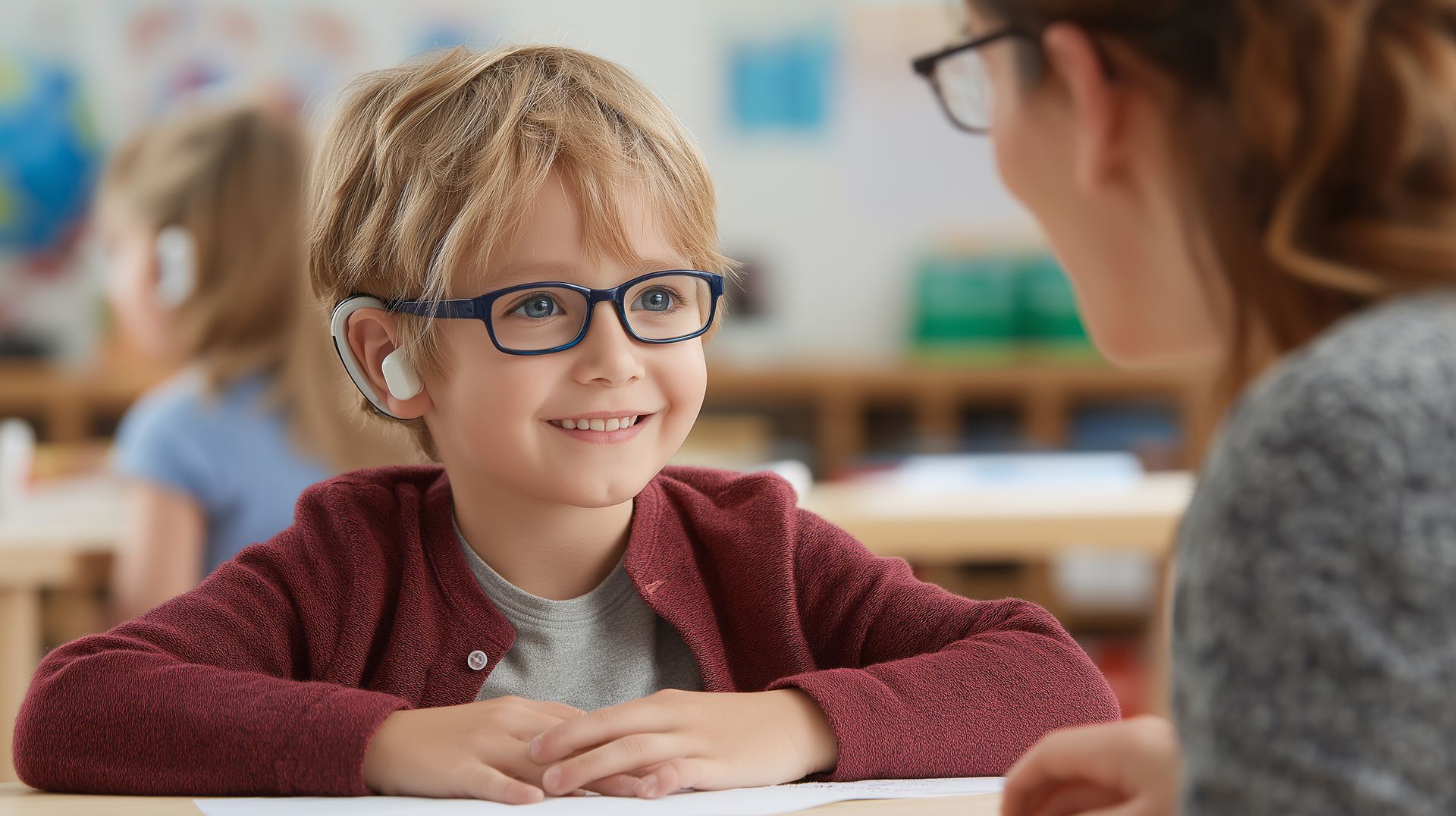 Enfant souriant portant des lunettes, assis à une table de classe, face à un enseignant au premier plan.