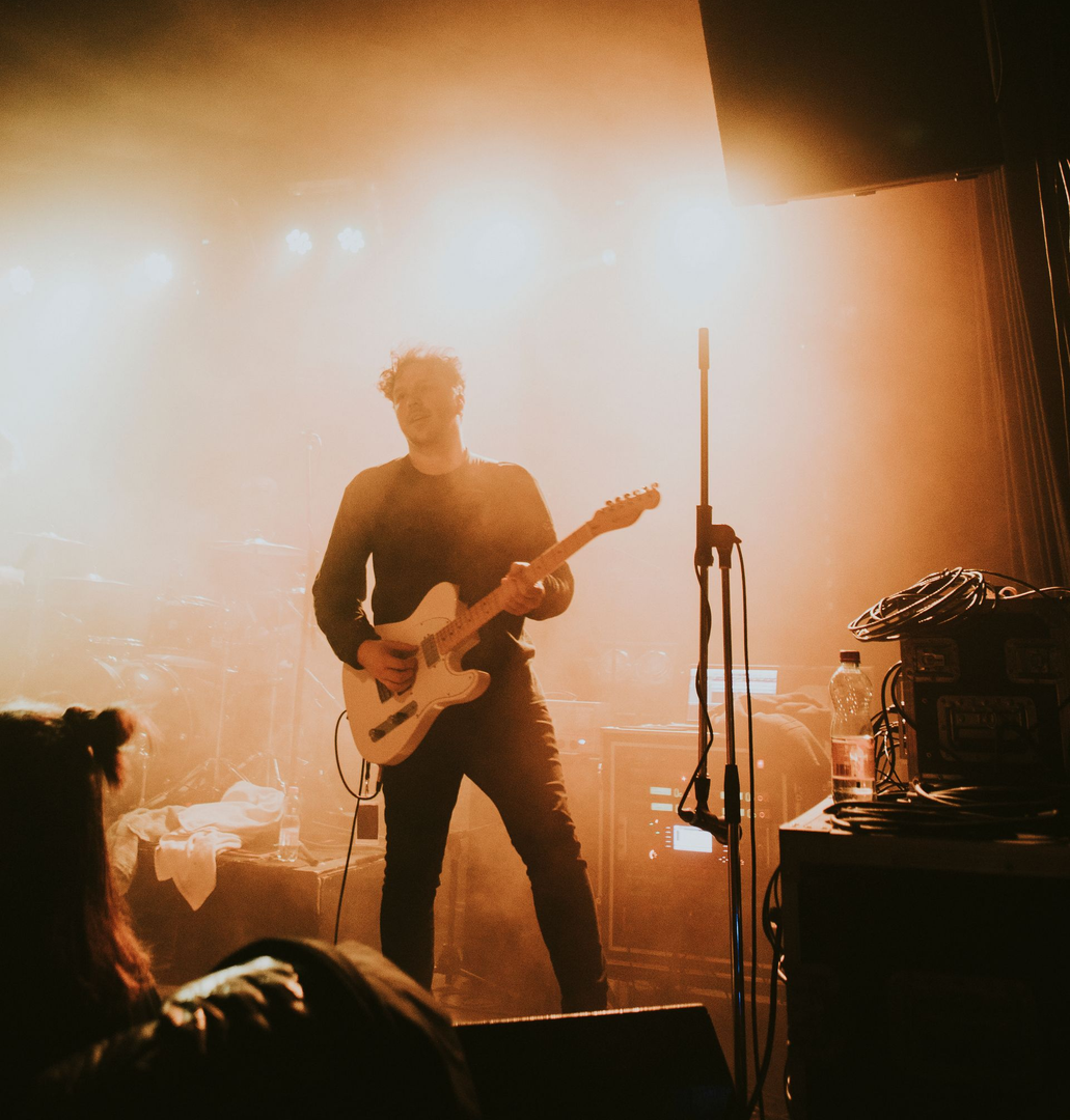 Un musicien joue de la guitare électrique sur une scène de concert éclairée d'une lumière orange enfumée.