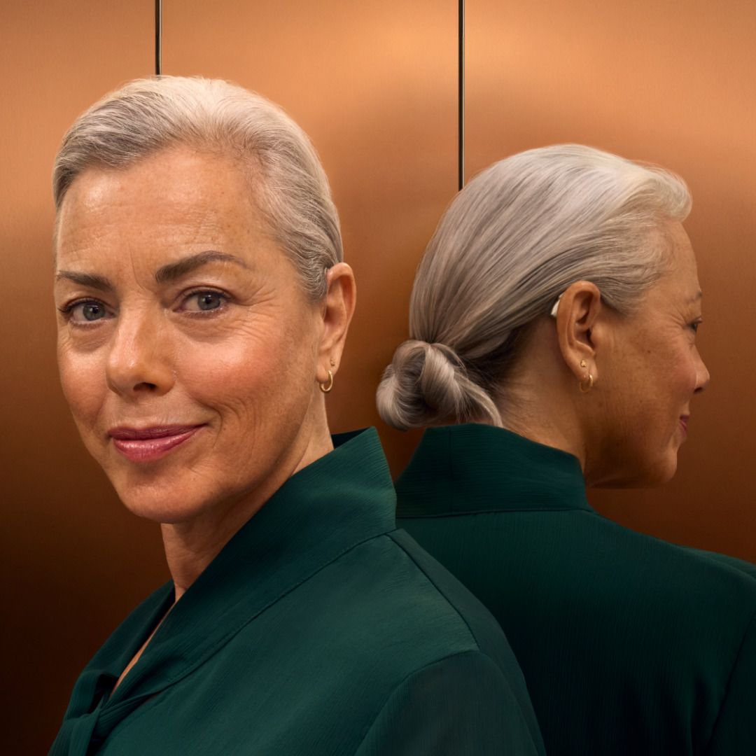 Femme portant un blazer vert foncé, photographiée de face et de dos devant une paroi d’ascenseur aux tons chauds.