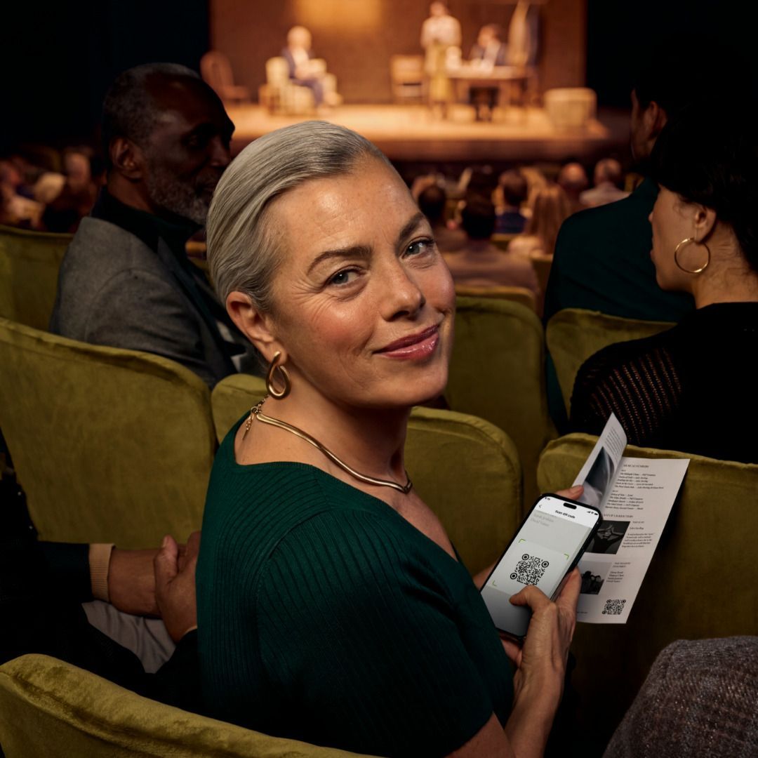 Personne souriante vêtue d’un haut vert, assise dans une salle de théâtre, tenant un programme .