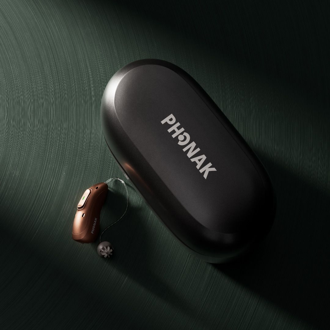 Étui noir pour appareil auditif Phonak, avec un appareil auditif posé à côté sur une surface vert foncé.