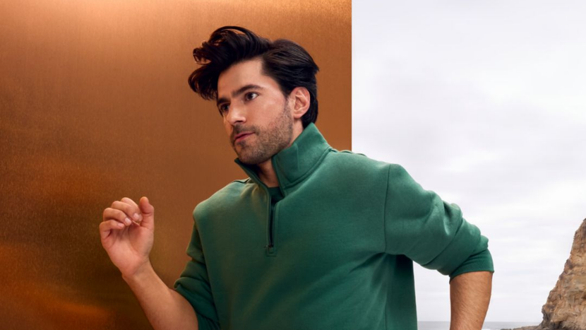 Un homme portant un pull vert zippé pose devant un fond brun chaud et blanc.