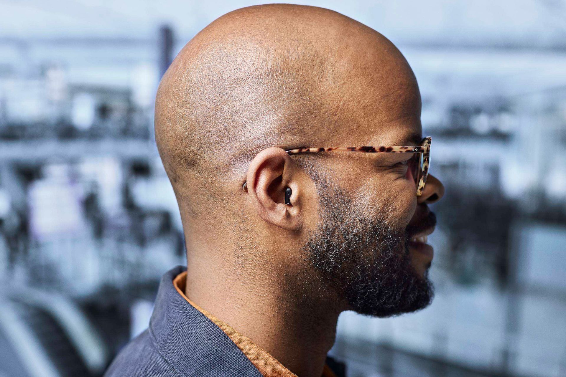 Portrait de profil d’un homme chauve portant des lunettes et une barbe, vêtu d’une chemise bleue sur fond extérieur flou.