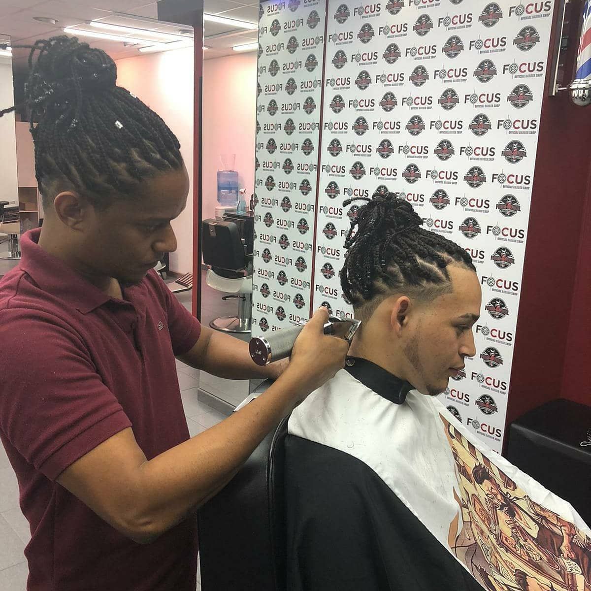 Barbero cortando el pelo en un salón. El cliente está sentado, con una capa. El logo de