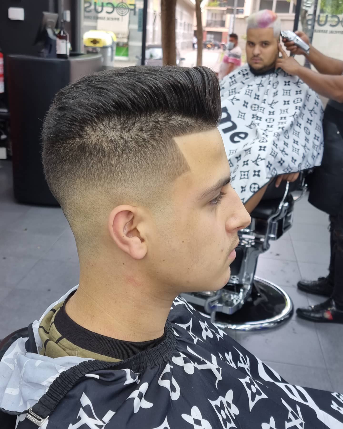 Un hombre con un corte de pelo pompadour en una barbería. Al fondo, otra persona se está cortando el pelo.