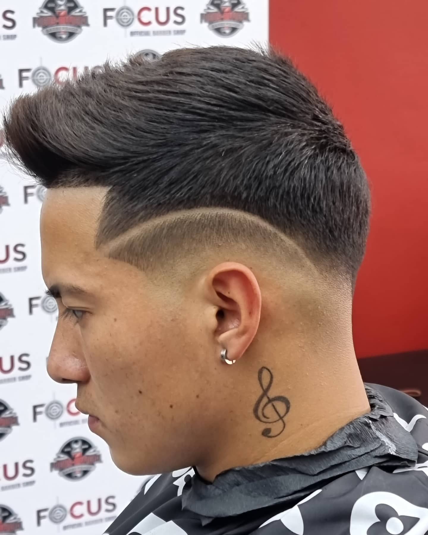 Hombre con corte de pelo descolorido y diseño lateral, vistiendo una capa estampada, con un tatuaje de clave de sol.