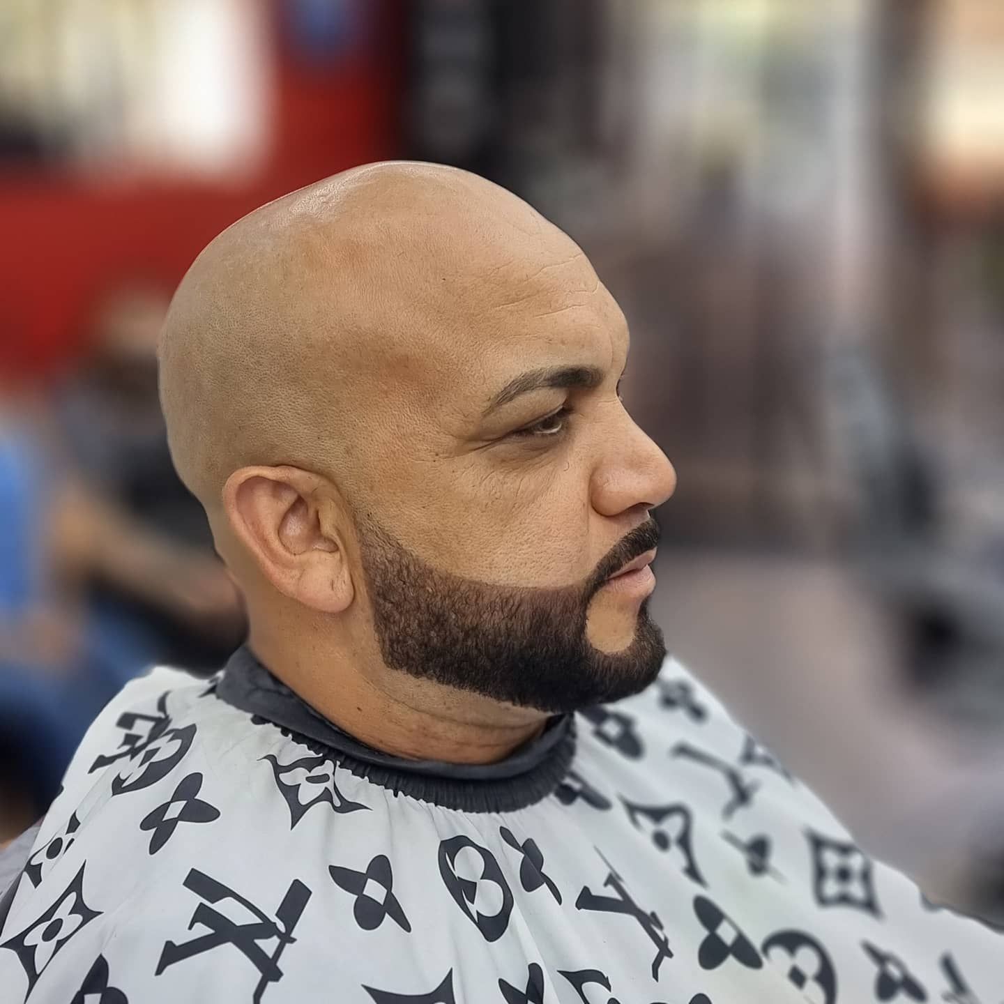 Hombre calvo con barba y cabello recortados, vistiendo una capa de barbero, mirando hacia la derecha, con una tienda de fondo.