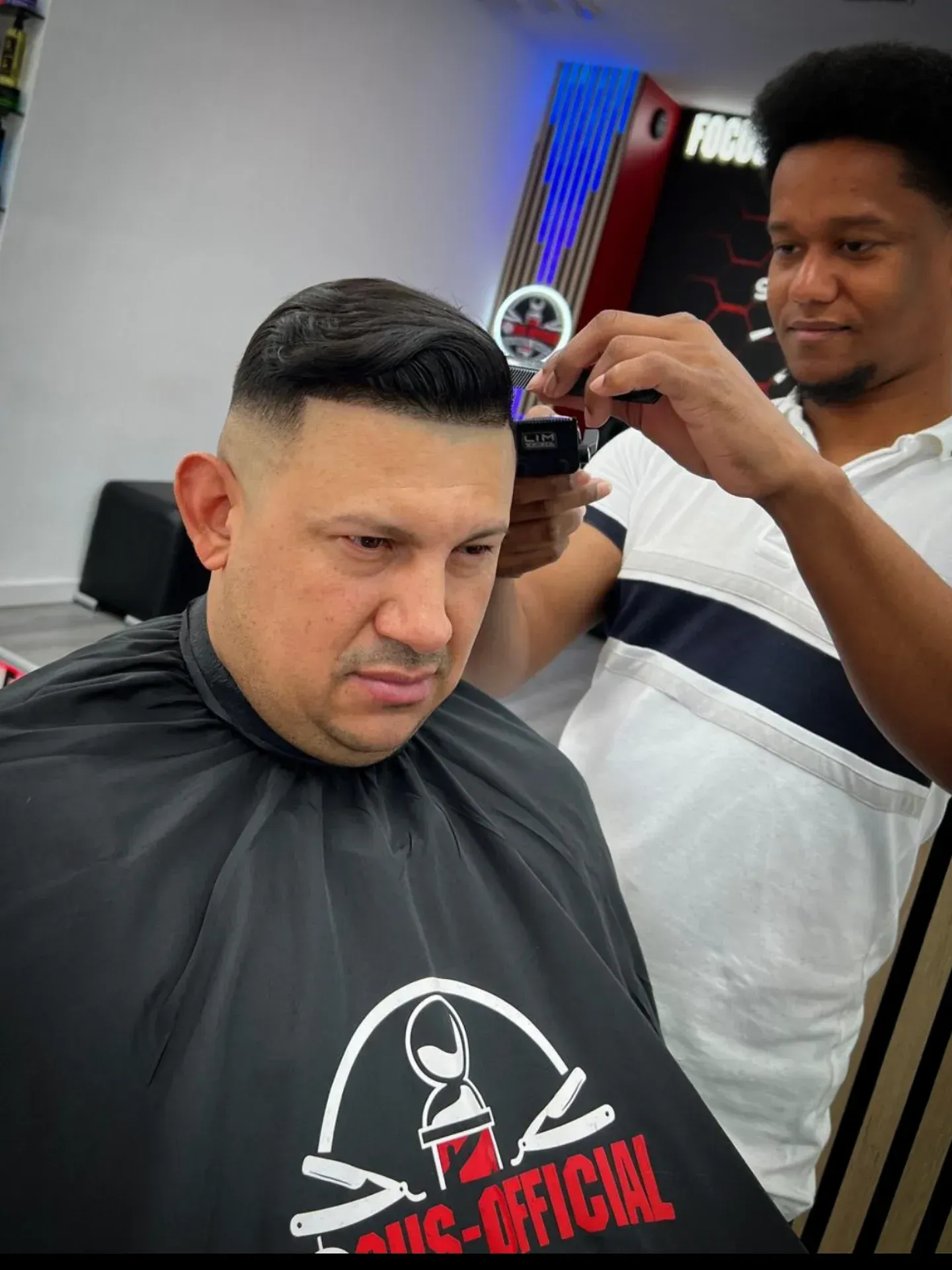 Un barbero corta el pelo a un cliente en una barbería. El cliente lleva una capa y el barbero usa una maquinilla.