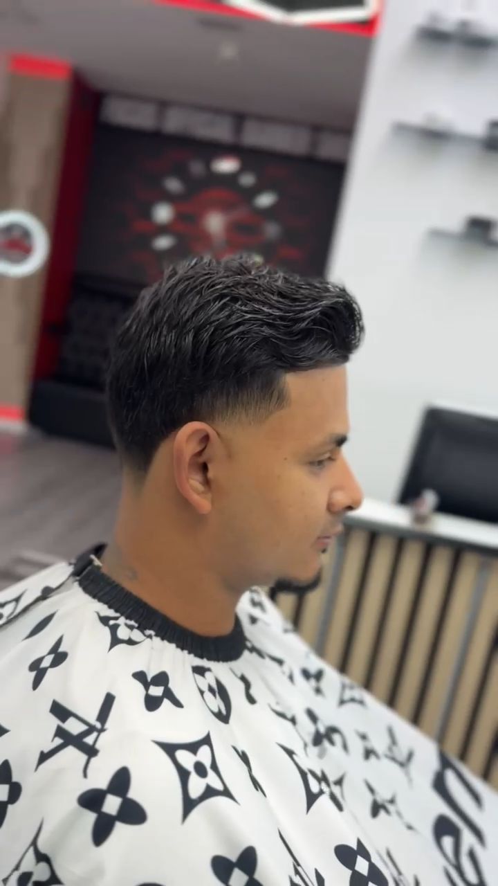 Hombre con un corte de pelo descolorido en una barbería, vistiendo una capa estampada, mirando hacia un lado.