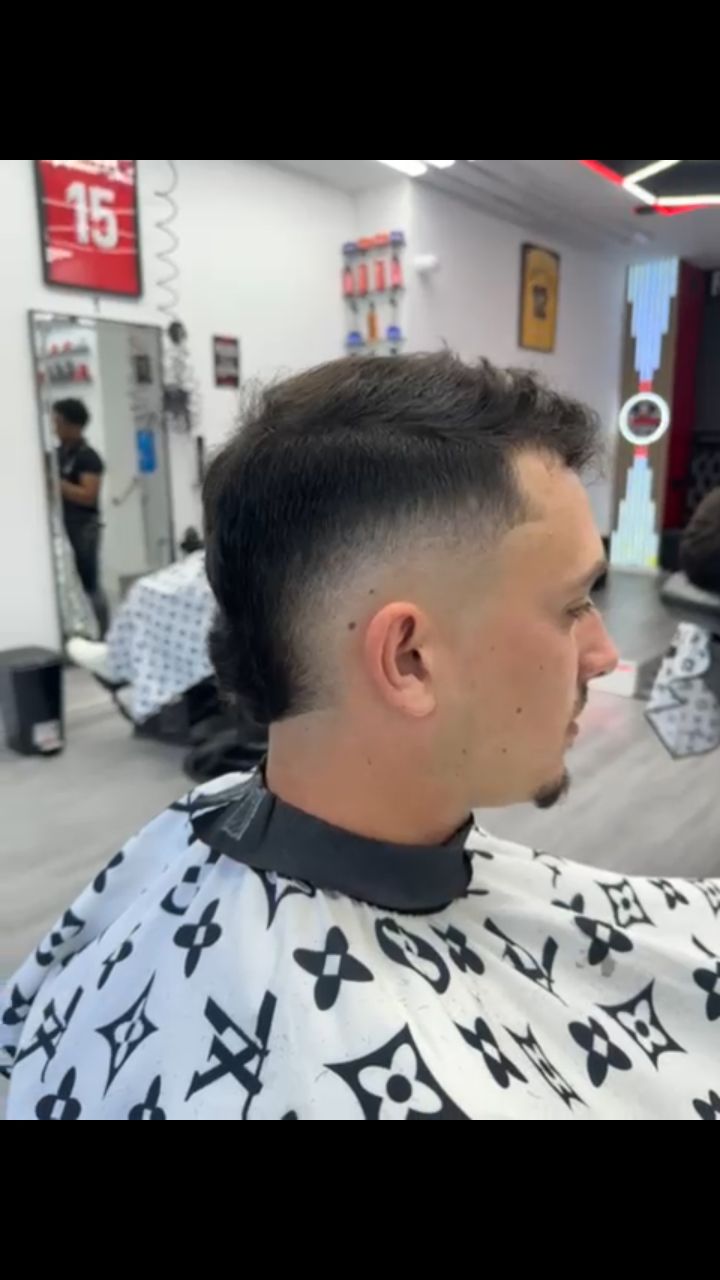Hombre con un corte de pelo descolorido en una barbería, vistiendo una capa con un diseño; mirando hacia la derecha.
