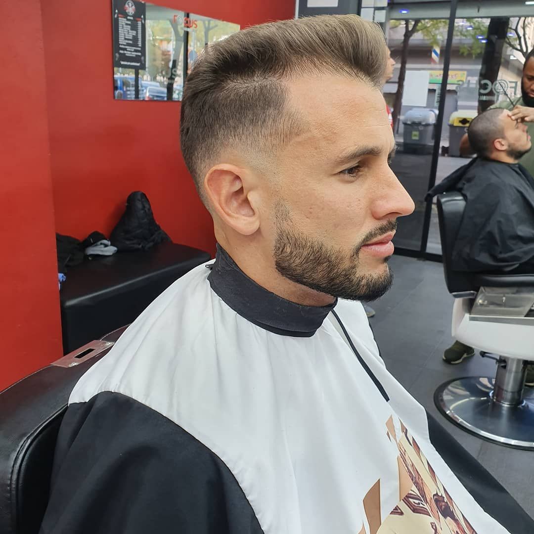 Hombre cortándose el pelo en una barbería; piel desvanecida, barba recortada, sentado en una silla, vistiendo una capa de barbero.