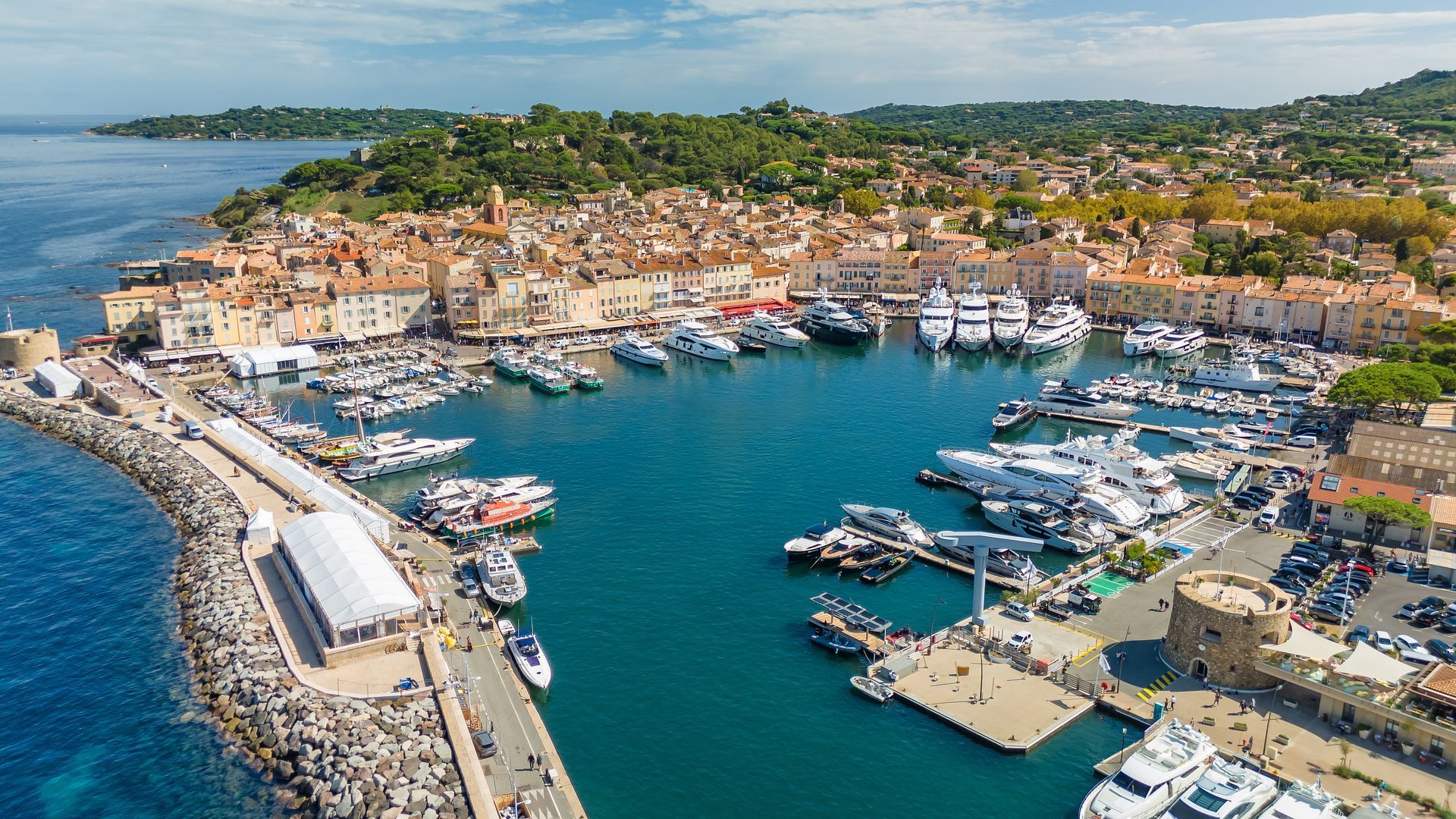Vue aérienne d'un port côtier avec des bateaux, des bâtiments en pierre et une eau turquoise le long d'une ville littorale