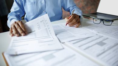 Une personne examine des documents et prend des notes à un bureau entouré de documents et de lunettes.