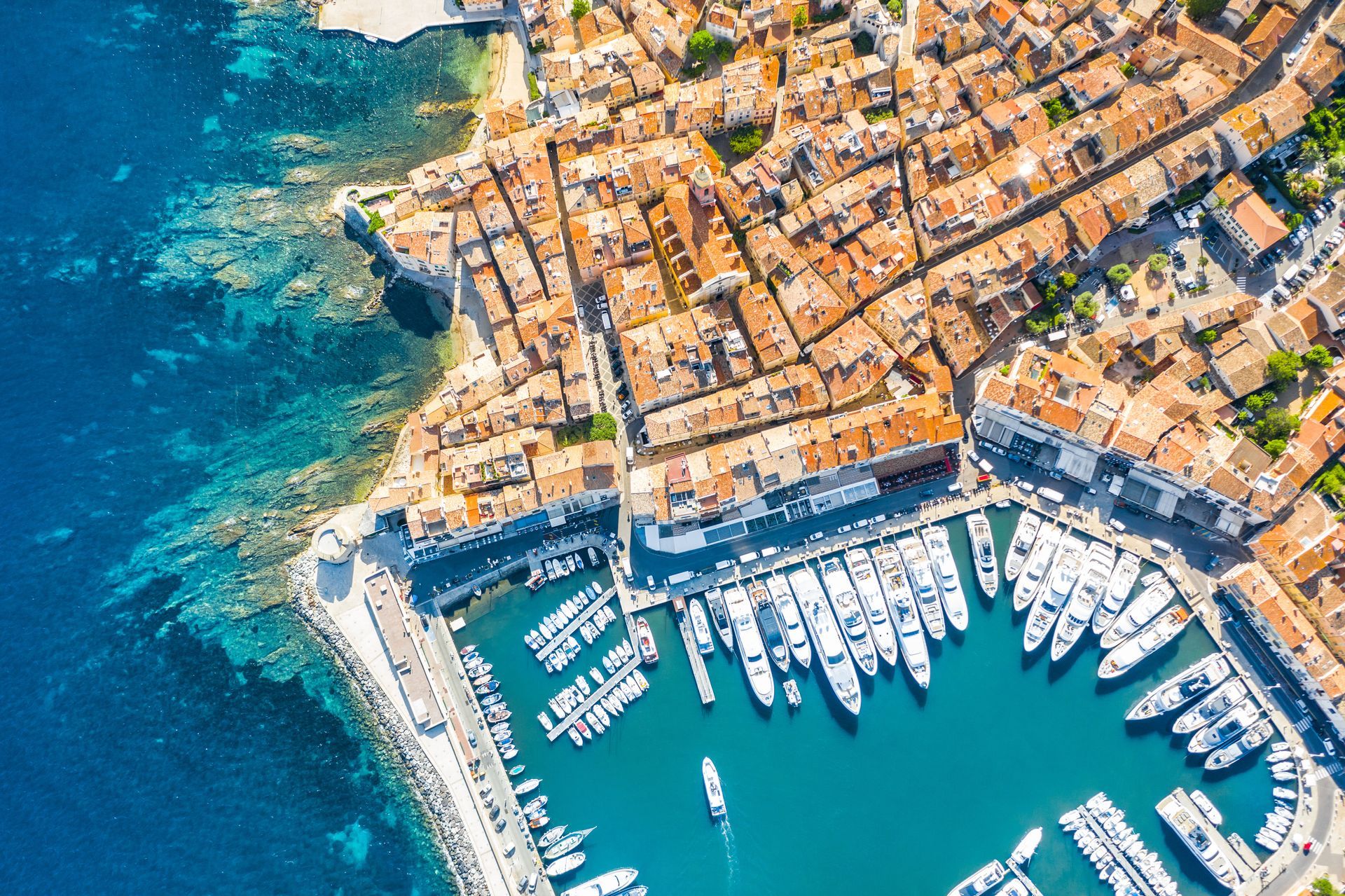 Vue aérienne d'un port de plaisance côtier avec des voiliers, une eau turquoise et une ville balnéaire densément peuplée.