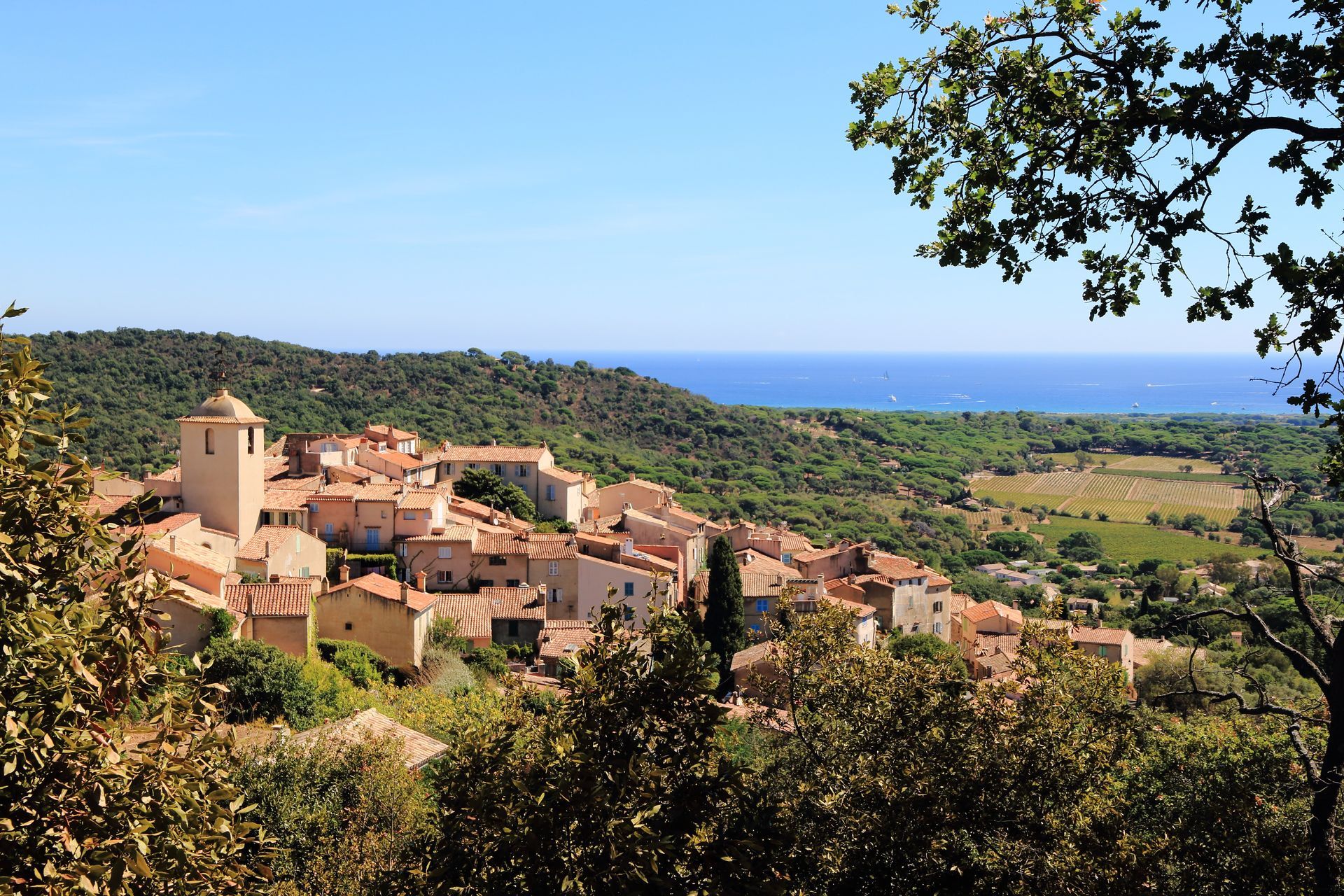Village perché sur une colline, avec ses bâtiments en terre cuite surplombant les champs verdoyants et la mer sous un ciel bleu.