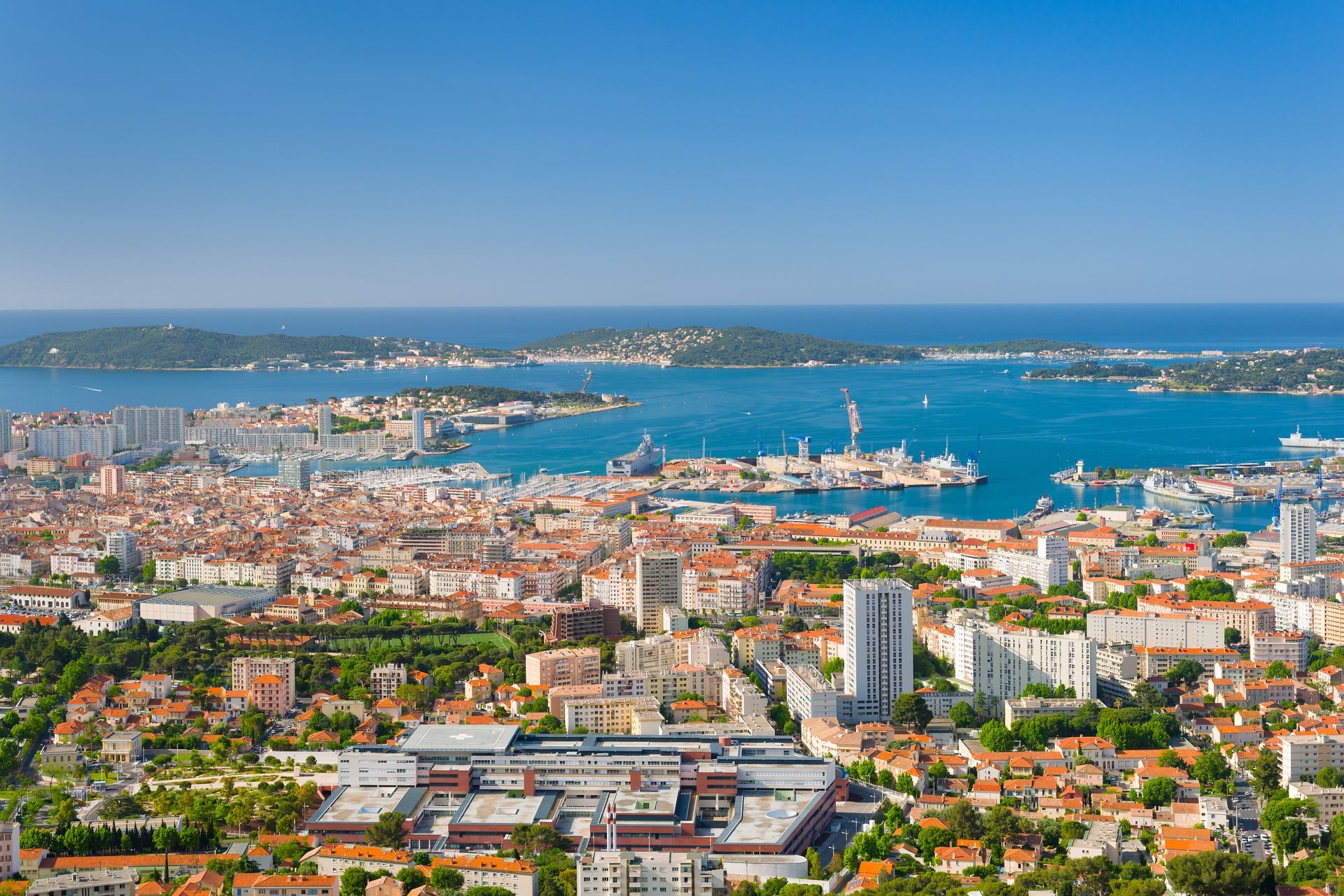 Vue panoramique d'une ville côtière et de son port sous un ciel bleu limpide, avec la mer, des bâtiments et des bateaux.