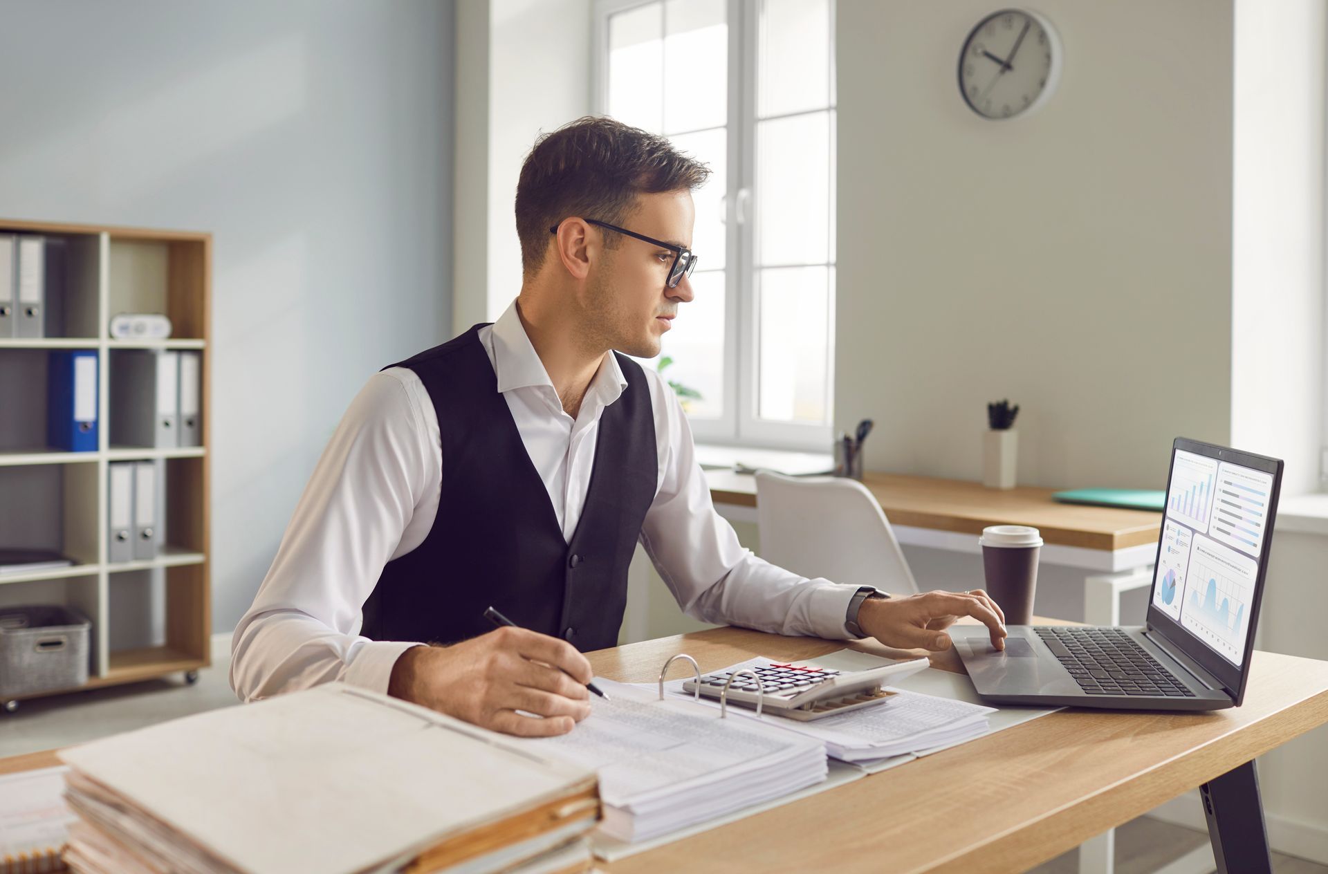 Homme portant des lunettes, un gilet, travaillant au bureau avec un ordinateur portable et une calculatrice, dans un environnement de bureau.