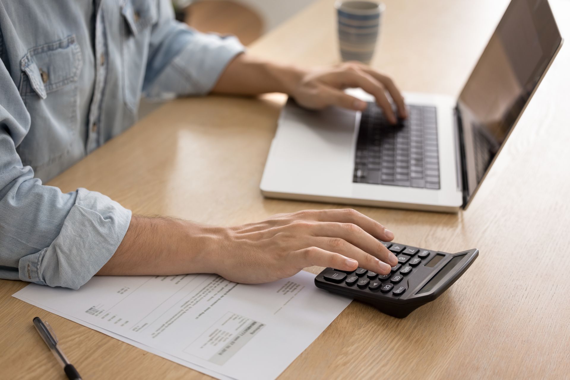 Une personne calcule avec une calculatrice tout en utilisant un ordinateur portable et en examinant un document à un bureau.