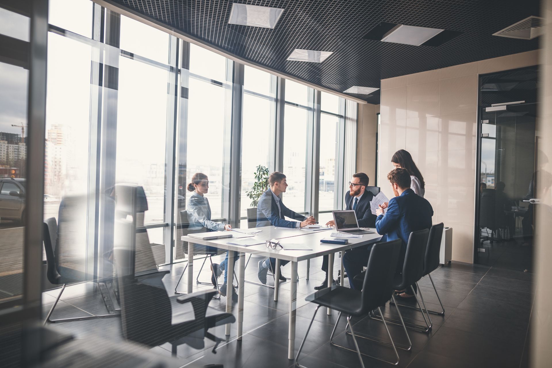 Une réunion d'affaires dans un bureau moderne, avec des personnes assises autour d'une table. La lumière du soleil filtre à travers de grandes fenêtres.