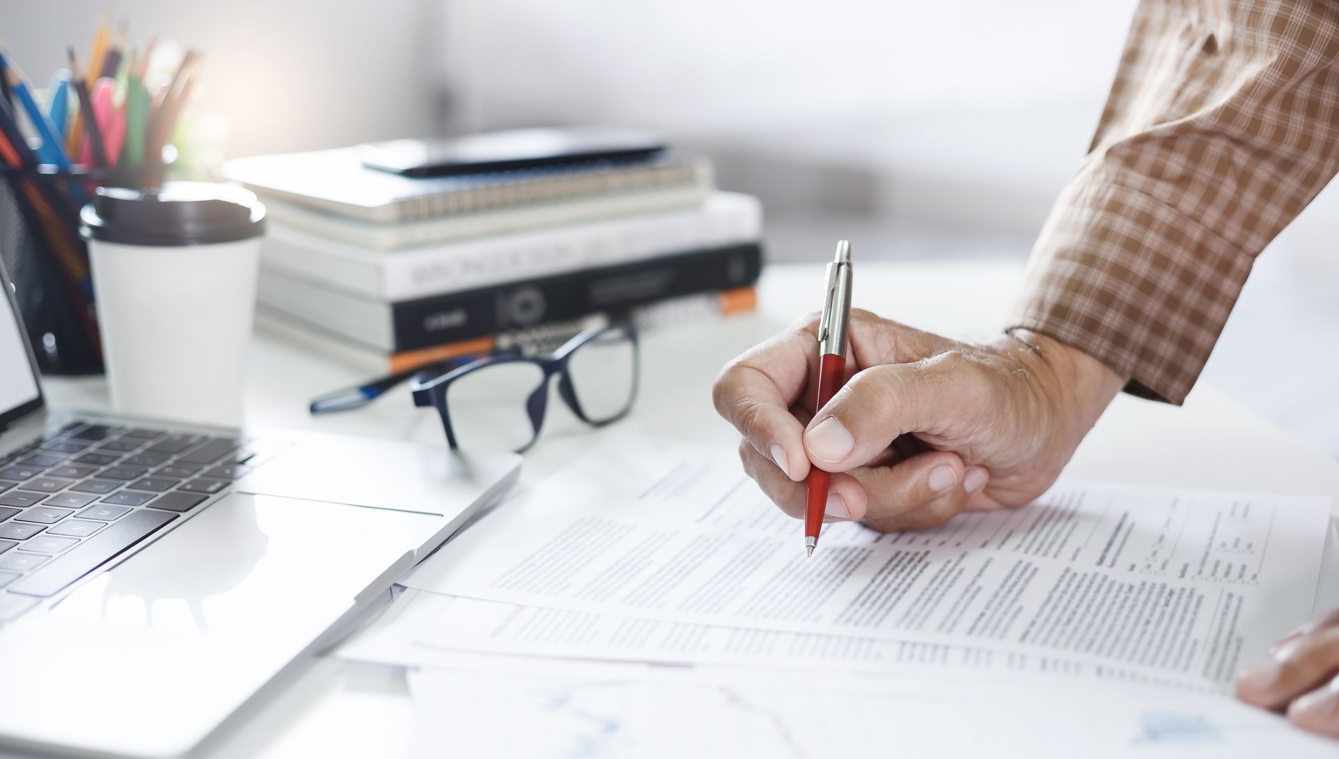 Personne signant des documents à un bureau avec un ordinateur portable, des lunettes, des livres et une tasse à café.
