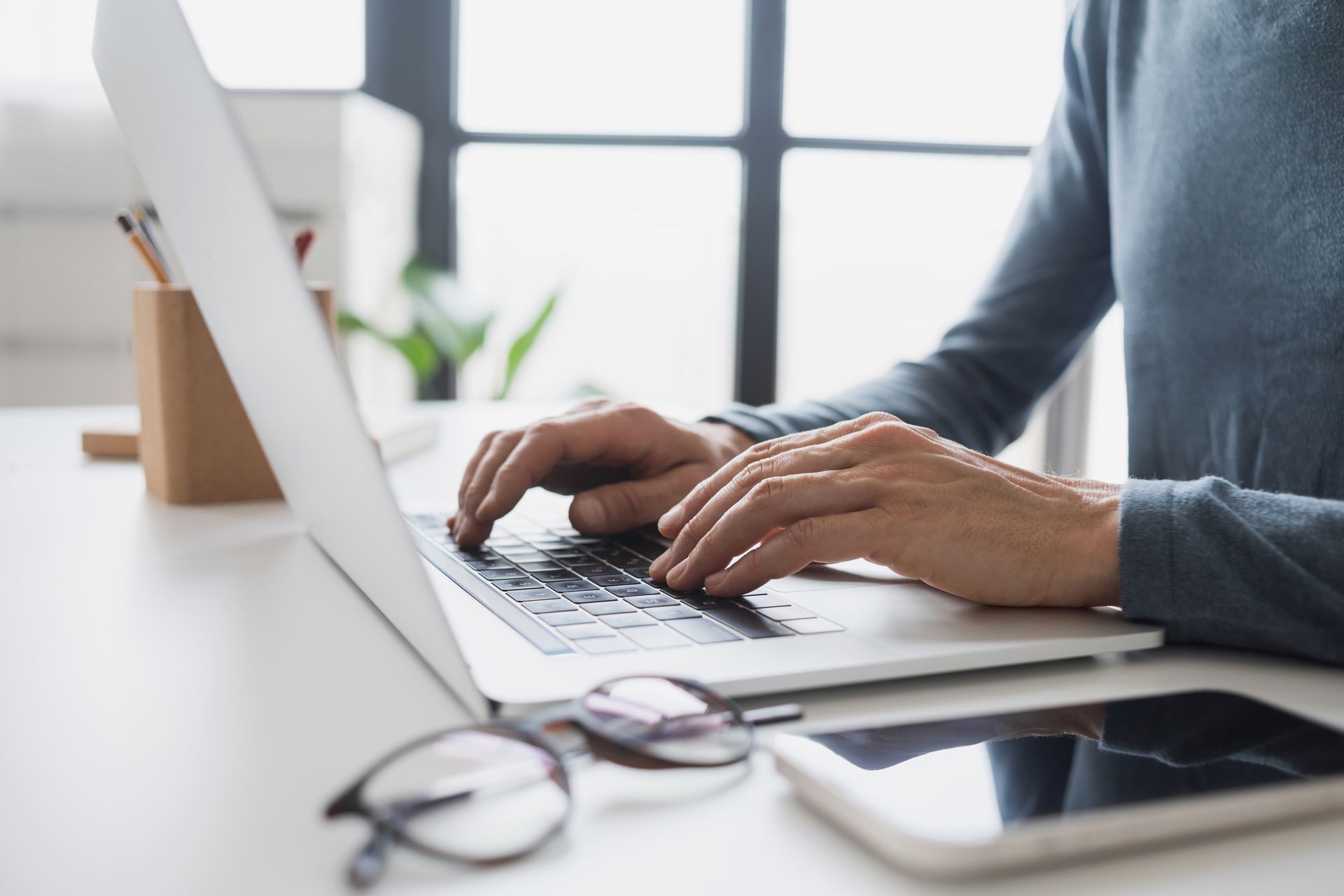 Personne tapant sur son ordinateur portable à un bureau blanc, lunettes et téléphone au premier plan. Fenêtre en arrière-plan.