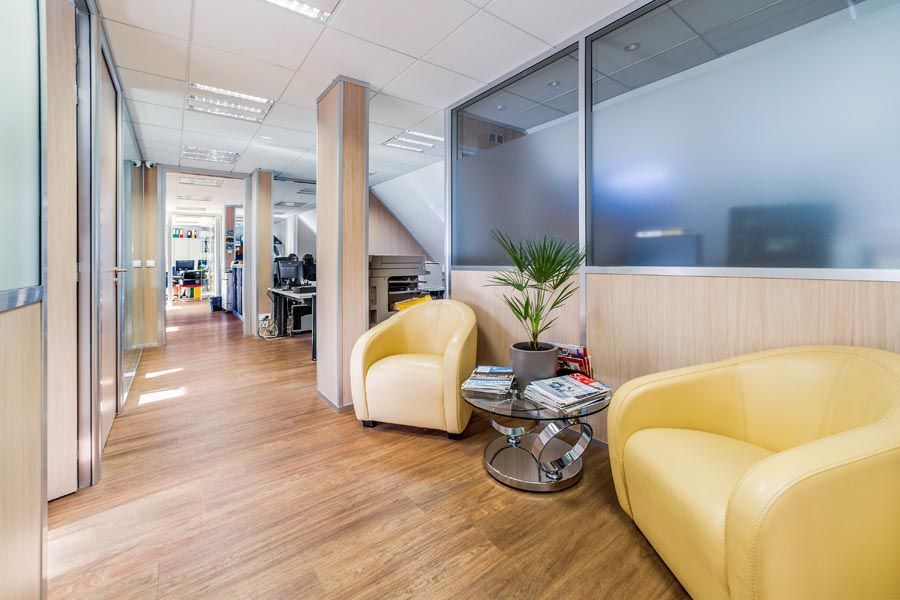 Couloir de bureau avec fauteuils jaunes, plante et fenêtres en verre dépoli.