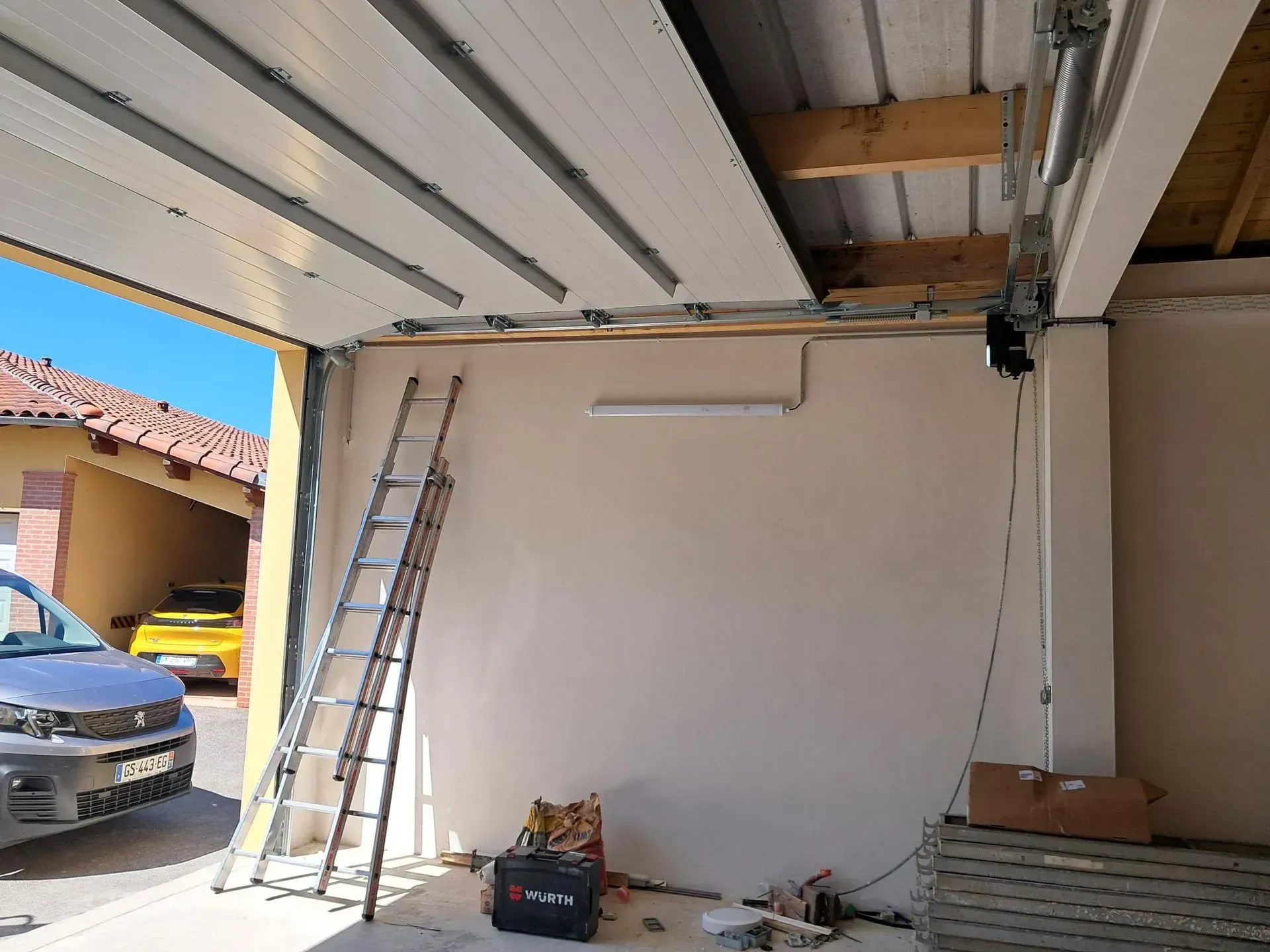 Intérieur d'un garage avec poutres apparentes, échelle, outils et voiture garée visible de l'extérieur.