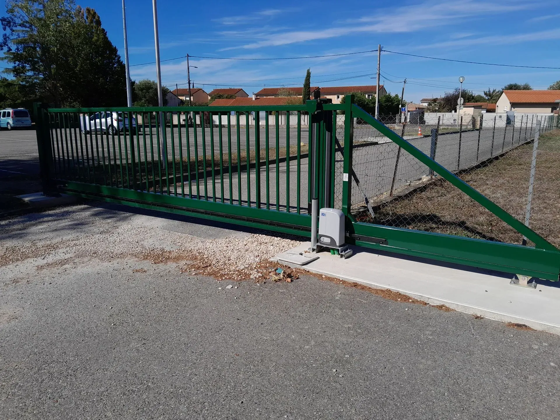 Portail d'allée en métal vert au bord d'un chemin de gravier, avec un pilier blanc et une cour clôturée sous un ciel dégagé